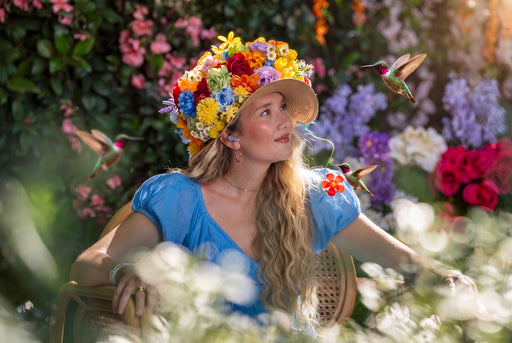A woman in a vibrant blue dress sits in a garden chair surrounded by colorful flowers and hummingbirds, wearing a large floral crown hat.