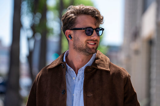 A man wearing black sunglasses with a thin metal frame and a brown suede jacket over a light blue button-down shirt, walking outdoors on a sunny day.