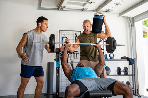 Three men in a home garage gym setting. One man is lying on a bench press, lifting a barbell, while another man stands behind him holding up a black speaker as a distraction.