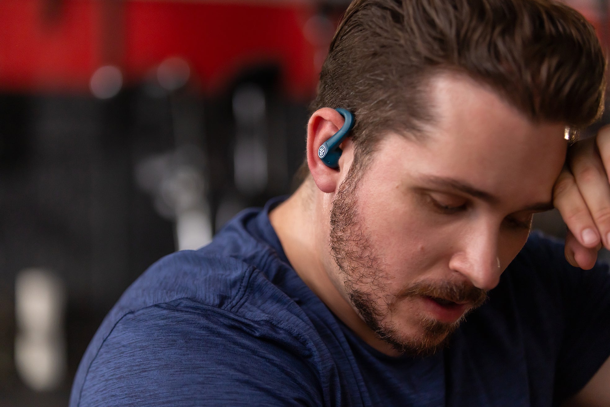 A close-up lifestyle shot of a man in a gym wearing a blue, ear-curved wireless earbud.