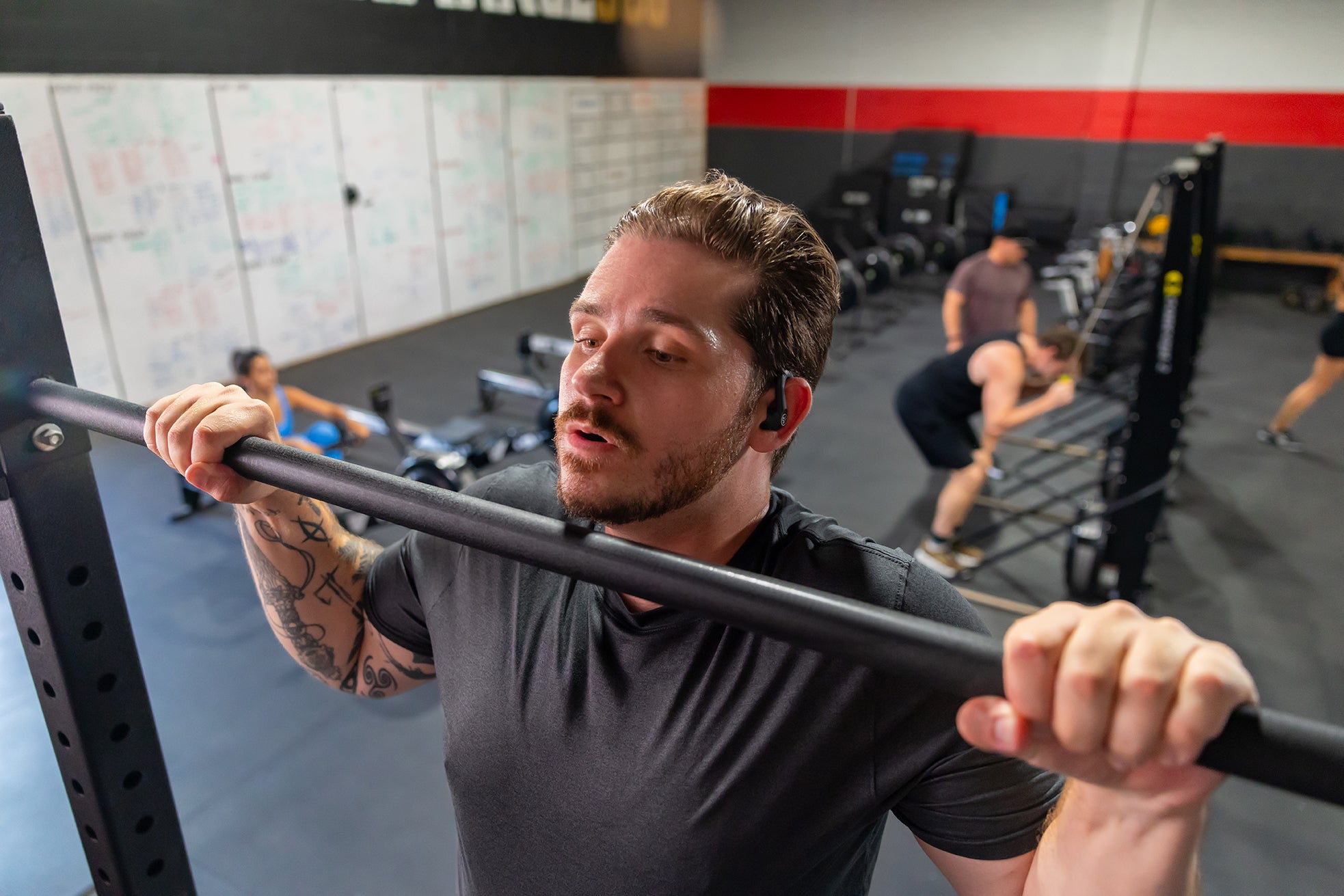 A man in a gym setting, wearing a dark gray t-shirt and holding a black barbell on his shoulders. Other gym-goers and exercise equipment are visible in the background.