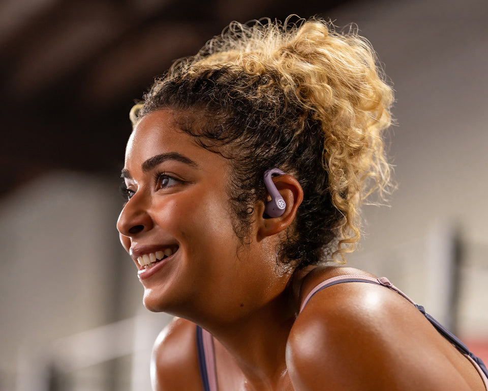 A close-up lifestyle shot of a woman with curly blonde hair wearing a purple wireless earbud in a gym setting.