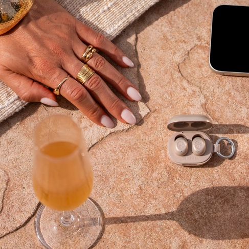 JBuds Mini ANC earbuds in the shade Gold Digger, laying on stone tile with a gold purse, gold heels, champagne, a phone, and a girl's hand wearing gold jewelry.