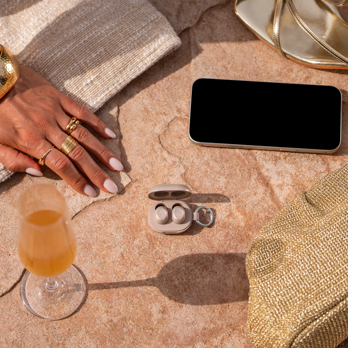 JBuds Mini ANC earbuds in the shade Gold Digger, laying on stone tile with a gold purse, gold heels, champagne, a phone, and a girl's hand wearing gold jewelry.