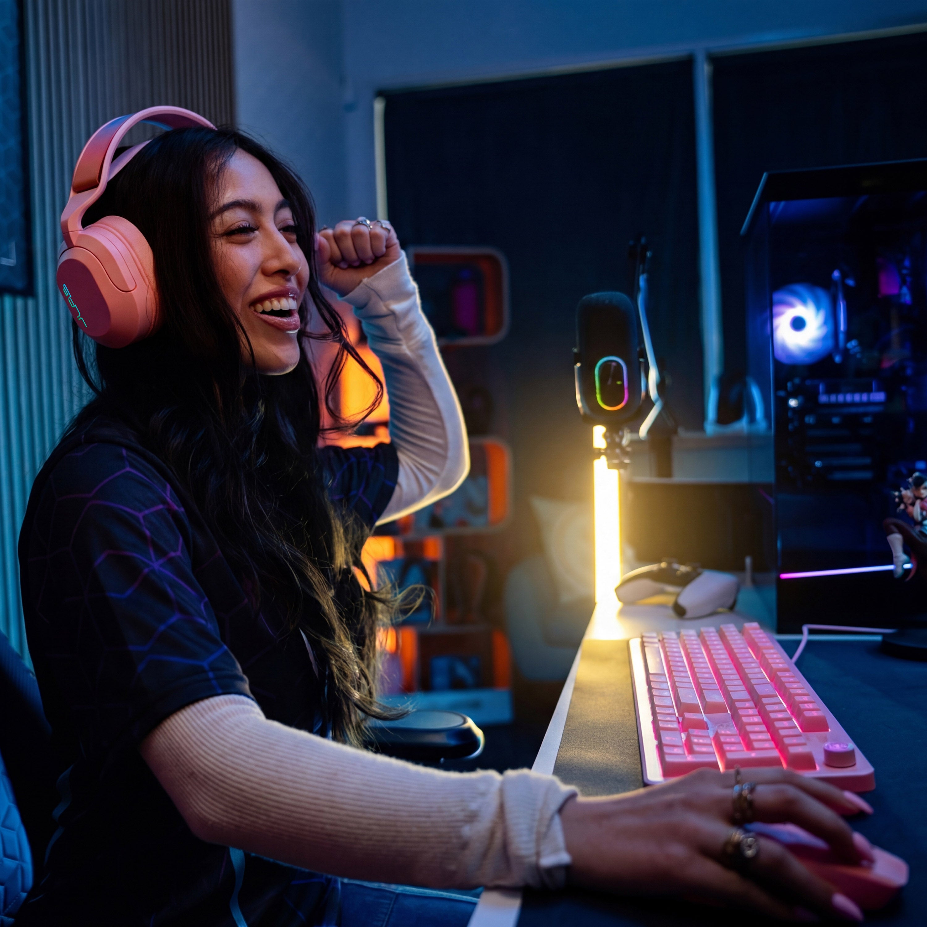 A woman wearing pink over-ear headphones sits at a desk in a dimly lit gaming room, smiling and laughing while using a pink keyboard and mouse. A microphone on a stand, computer tower, and ambient blue lighting are visible in the background.