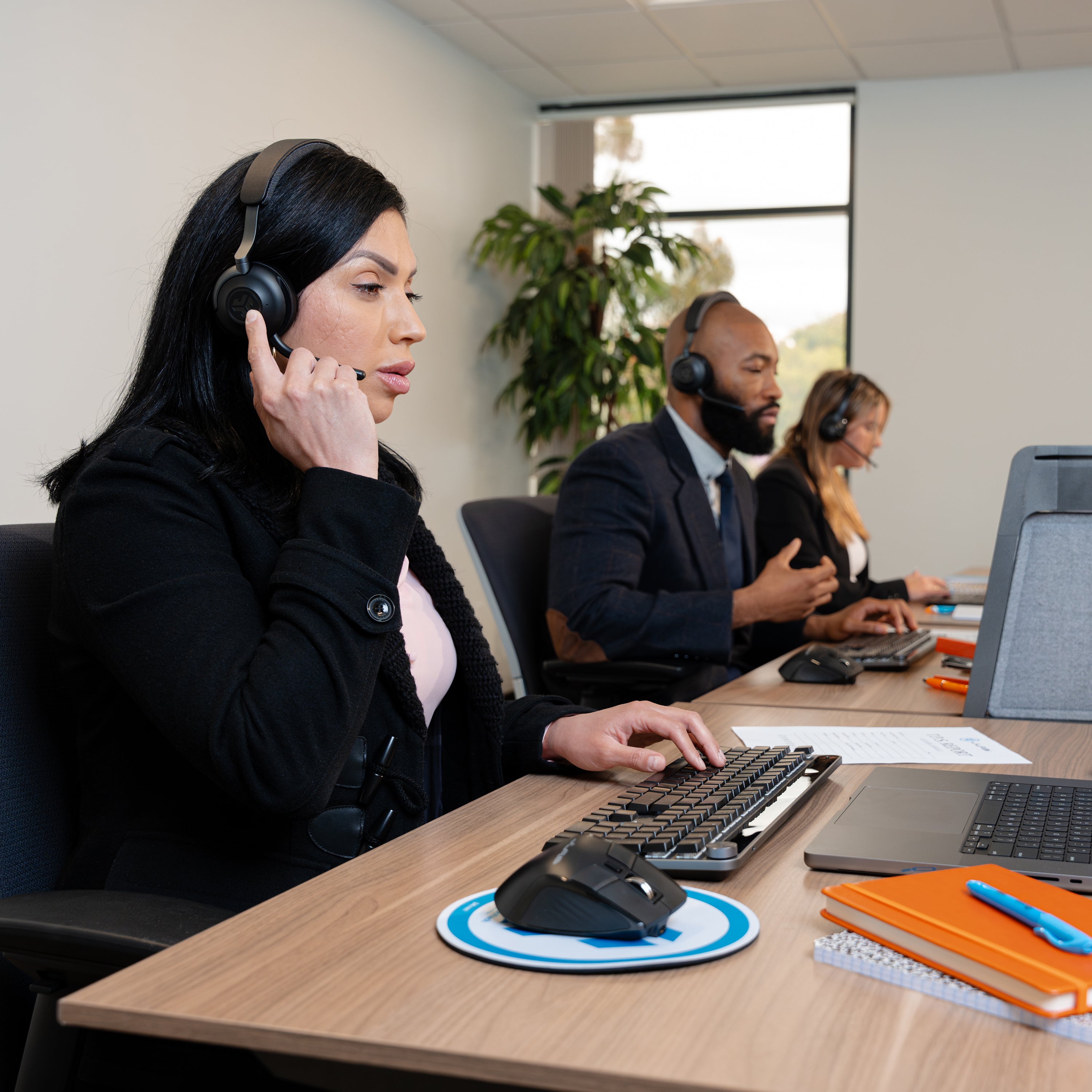 A woman in a black blazer wearing a headset sits at a desk using a computer in an office setting, with two other employees in the background.
