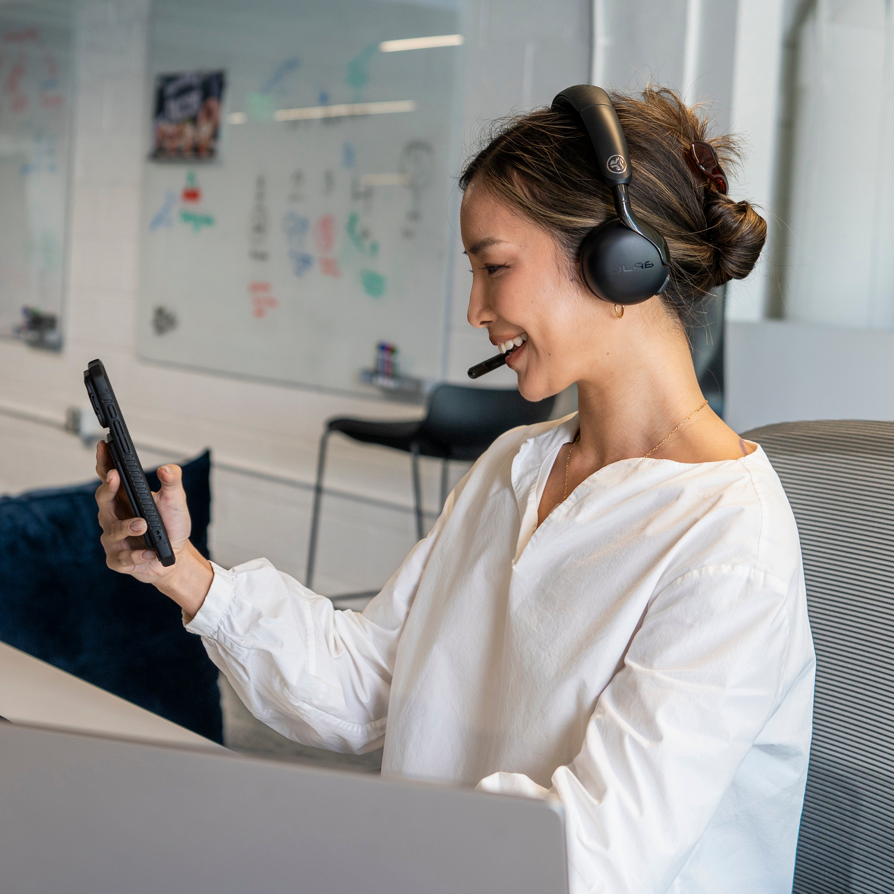 A woman in a white blouse wearing black over-ear headphones with a microphone sits at a desk, holding a smartphone in a bright modern office.