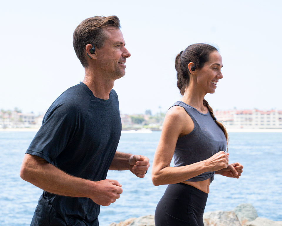 A man and a woman jogging outdoors by the water, both wearing wireless earbud-style fitness earpieces.