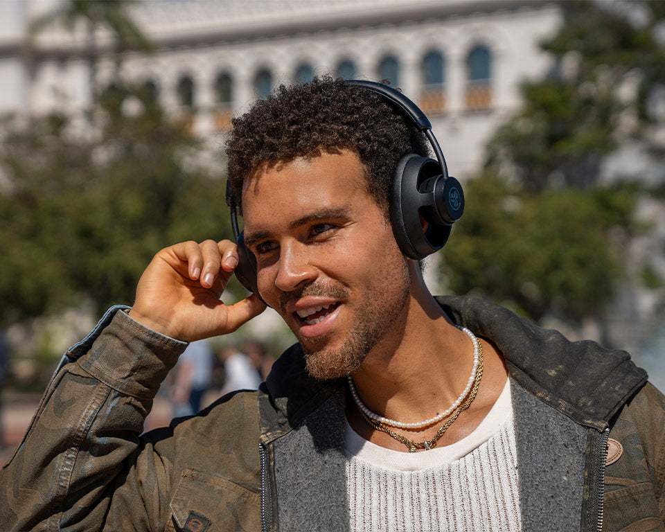 A man outdoors wearing black over-ear headphones and a brown jacket over a white shirt, with a blurred building and trees in the background.