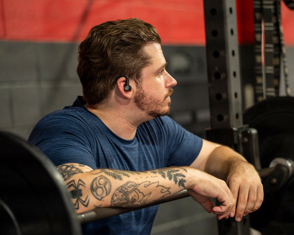 A man with tattoos in a gym setting, wearing a blue t-shirt and a black wireless earbud, leaning on a barbell rack.